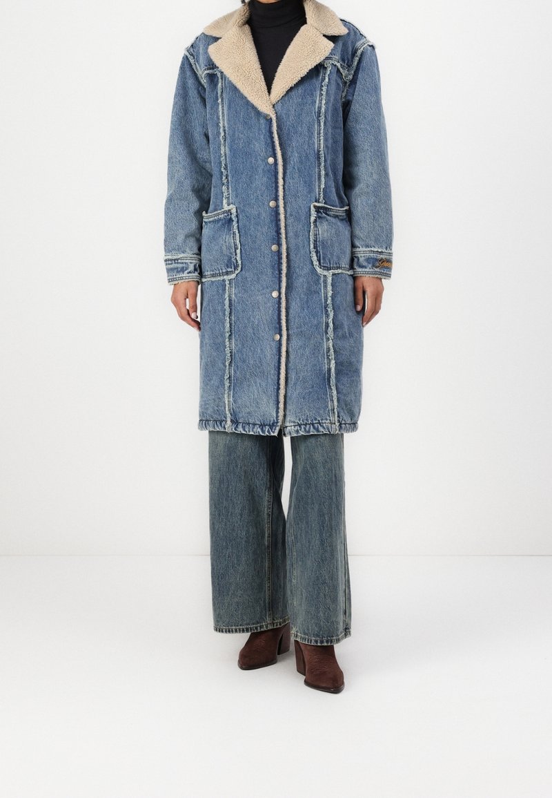 Manteau en denim long avec un col en fausse fourrure beige, fermeture à boutons sur le devant, deux grandes poches à rabat et détails de couture contrastants. Porté sur un jean large.