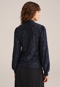 Marinblus med broderi och långa ärmar, med ett texturerat blommönster och en figurnära design, tillsammans med en veckad svart kjol.