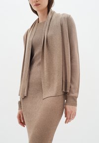Beige kavaj med öppen front, mjuk textur och långa ärmar, lagrad över en matchande ribbad klänning. Båda plaggen har en subtil glans.