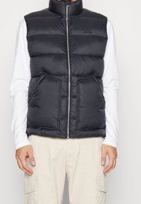 Svart quiltad ärmlös väst med framzipper och fickor, bärs över en vit långärmad skjorta och beige cargobyxor.