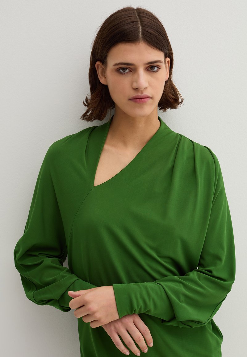Groen, langwerpig shirt met een V-hals, gedrapeerde stof, een lichte plooiing op de mouwen en een gladde textuur tegen een neutrale achtergrond.