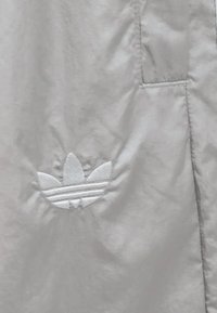 Ljusgrå vindjacka med en subtil textur och en broderad vit tre-stripes-logotyp nära fickan på vänster sida.