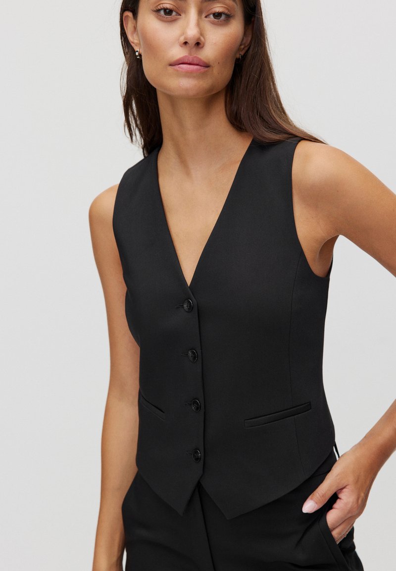 Zwarte vest met een diepe V-hals, zes knopen op de voorkant en een getailleerde pasvorm. Voorzien van twee zijzakken en een gladde stofstructuur.