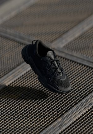 Sort atletiksneaker ophængt over et tekstureret metalristgulv med krydsmønster.