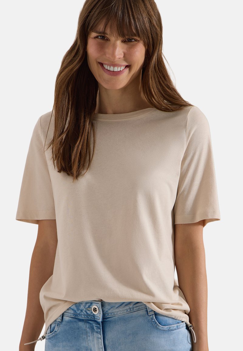 Femme souriante aux longs cheveux bruns portant un t-shirt beige à manches courtes et un jean bleu clair sur un fond uni.
