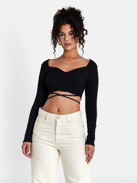 Black ribbed crop top with long sleeves and a heart-shaped neckline. Features crisscross detailing at the waist. Paired with light-colored pants.