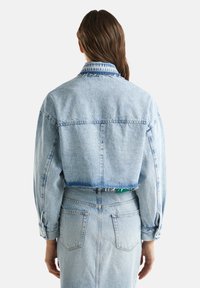 Beskuren ljusblå denimjacka med långa ärmar, fransig nederkant och en kontrasterande mönstrad krage. Har standard jackstygn och fickor.