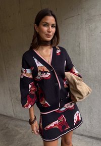 Γυναίκα που φοράει μαύρη ζακέτα και φούστα με πολύχρωμα αφηρημένα σχέδια, κρατώντας μπεζ αναδιπλωμένο φάκελο μπροστά σε φόντο από σκυρόδεμα.