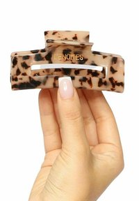 Sköldpaddsfrisyrklämma med glänsande yta, rektangulär form och guldfärgad "LENOTES"-gravyr. Hållen i en hand med neutral nagellack.