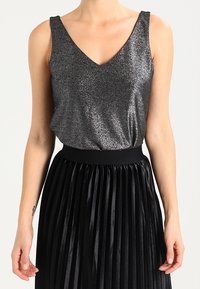 Silber schimmerndes Tanktop mit V-Ausschnitt, kombiniert mit einem hoch taillierten schwarzen Faltenrock. Der Rock hat eine strukturierte, glänzende Oberfläche.