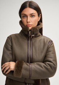 Bruine shearlingjas met een hoge kraag, bruine bontafwerking en donkere lederen accenten; voorzien van een rits aan de voorkant en zachte mouwen.