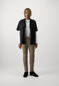 Short-sleeve black shirt with two pockets, layered over a white long-sleeve shirt; tan pants and black sneakers complete the outfit.