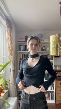 Juoda pintinė ilgomis rankovėmis palaidinė su V formos iškirpte ir chokero detale; derinama su aukštu liemeniu tamsiomis džinsais. Ant liemens matomi subtilūs tatuiruotės.
