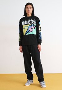 Person with shoulder-length black hair wearing black graphic long-sleeve shirt, black pants, and gray sneakers standing on yellow floor.