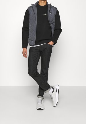 Hombre con sudadera negra, chaqueta sin mangas acolchada gris, vaqueros oscuros y zapatillas blancas, de pie contra un fondo liso con una mano en el bolsillo.