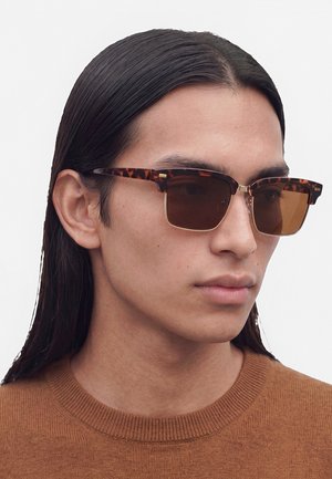 Personne aux longs cheveux foncés portant des lunettes de soleil carrées écaille de tortue et un pull en laine marron, regardant légèrement sur le côté.