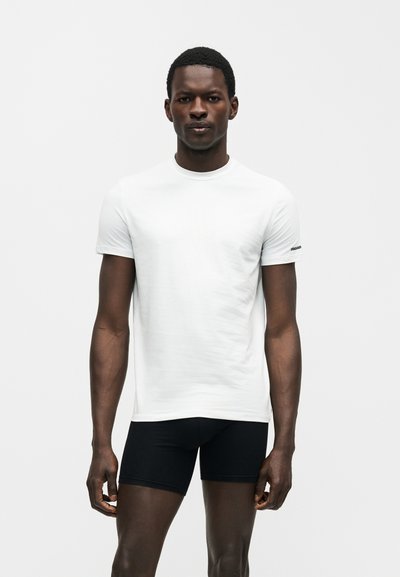 Άνδρας με σκουρόχρωμο δέρμα που φορά απλό λευκό κοντομάνικο μπλουζάκι και μαύρα στενά σορτς, στέκεται μπροστά σε λευκό φόντο.