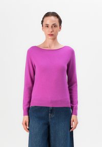 Jasnoróżowy sweter z szerokim, płaskim dekoltem i długimi rękawami. Wykonany z miękkiego materiału z prążkowanym wykończeniem, zestawiony z ciemnymi dżinsami.