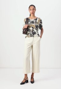Blusa a maniche corte con motivo floreale nei colori crema, nero e marrone, abbinata a pantaloni larghi crema e scarpe nere con tacco. Borsa marrone.