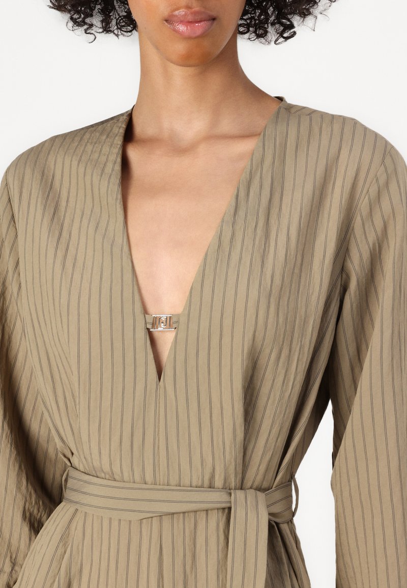 Beige gestreiftes Kleid mit tiefem V-Ausschnitt und weiten Ärmeln. Besitzt einen silbernen Akzent vorne und einen passenden Gürtel in der Taille.