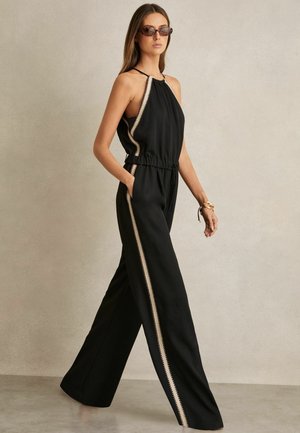 Kvinde iført en sort ærmeløs jumpsuit med beige kant, vide ben, solbriller og armbånd, stående mod en ensfarvet beige baggrund.