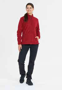 Röd fleecjacka med hög krage, främre blixtlås och sidofickor, tillsammans med svarta byxor och svarta vandringsskor. Slät textur.