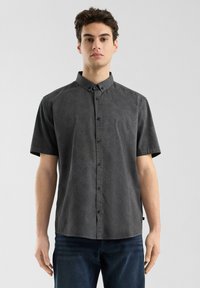 Camisa de manga corta con botones en tejido gris oscuro con un patrón sutil, presenta un cuello y un corte recto. Llevada con unos vaqueros oscuros.