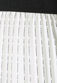 Falda blanca con pliegues que presenta una cinturilla elástica negra con texto en relieve. La tela tiene patrones verticales en texto negro por toda la superficie.