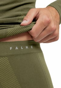 Tirant sur un vêtement seamless vert olive laissant apparaître la taille avec le logo "FALKE" en lettres blanches.