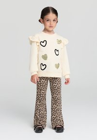 Maglione crema con motivi a cuore neri e luccicanti, maniche a volant. Indossato con pantaloni svasati beige con stampa leopardata e scarpe nere.