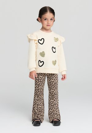 Maglione crema con motivi a cuore neri e luccicanti, maniche a volant. Indossato con pantaloni svasati beige con stampa leopardata e scarpe nere.