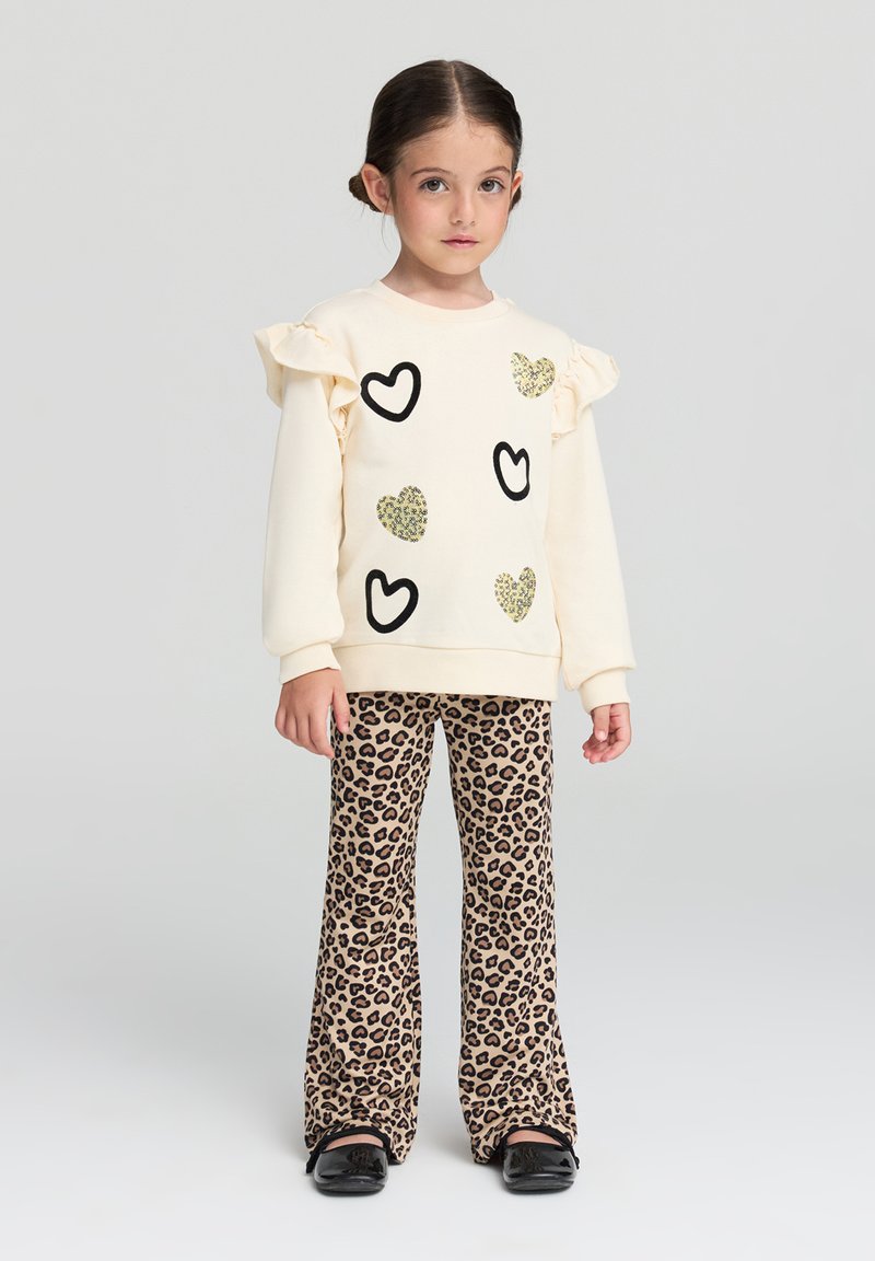 Maglione crema con motivi a cuore neri e luccicanti, maniche a volant. Indossato con pantaloni svasati beige con stampa leopardata e scarpe nere.