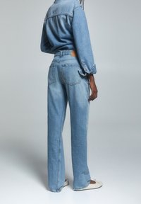 Persona vestida con una camisa de mezclilla azul clara y jeans, con bailarinas blancas, de pie contra un fondo gris claro y liso.