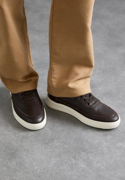 Brown leather sneakers with textured finish, white rubber sole, and brown accents. Worn with tan pants on a gray surface.