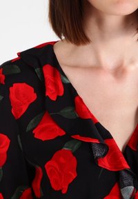 Sort bluse med rødt rosemønster, prydet med en flæse ved halsudskæringen og korte ærmer. Stoffet ser let og glat ud.