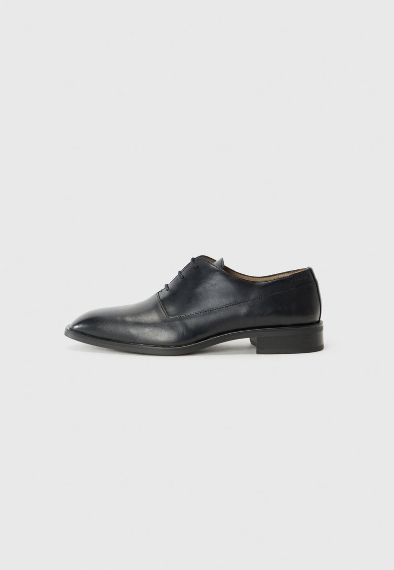 Zwarte leren dresschoenen met een puntige neus, vetersluiting en een lage hak, gepresenteerd tegen een eenvoudige witte achtergrond.
