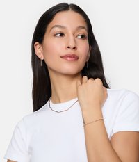 Roze gouden sieradenset, waaronder een ketting en armband, met langwerpige, geometrische schakels en kleine kralen, gedragen met een effen witte shirt.