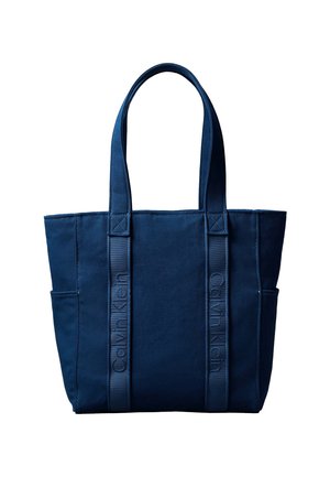 Torba tote Calvin Klein v mornarsko modri barvi z dvema dolgima ročajema, stranskimi žepi in navpičnimi trakovi z logotipom na ravni beli podlagi.
