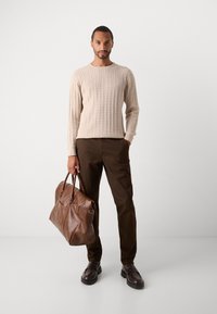 Man in beige cable-knit sweater and brown pants holding a large brown leather duffel bag, standing against a plain white background.