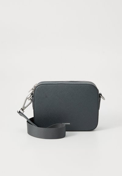 Sort rektangulær crossbody-taske med en struktureret overflade, sølvhardware og justerbar skulderrem. Indeholder en lyn lås og brandlogo foran.