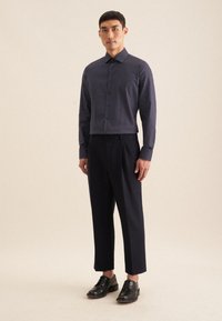 Marineblauwe blouse met witte stippen, lange mouwen, gecombineerd met donkere op maat gemaakte broeken en zwarte veterschoenen, staande op een neutrale achtergrond.