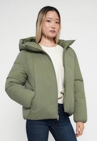 Giacca puffer verde oliva con cappuccio, chiusura con zip e tasche laterali. Tessuto liscio con una finitura leggermente lucida. Design casual.