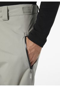 Lichtgrijze broek met een gladde textuur, voorzien van een ritszak en een stoffen patch op de tailleband. Zwarte overshirt met lange mouwen zichtbaar.