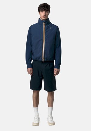 Uomo con giacca blu navy con zip, pantaloncini neri fino al ginocchio, calzini bianchi e scarpe da ginnastica bianche, in piedi davanti a uno sfondo uniforme.