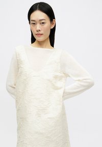 Robe couleur crème avec une surface texturée, présentant un décolleté en V profond, superposée à un haut en maille légère et transparente à manches longues.