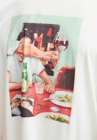 T-shirt blanc présentant une illustration colorée de mains portant un verre de vin, entourée d'une mise en scène de table avec de la nourriture et des boissons.