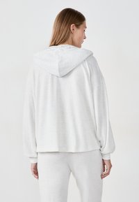 Sudadera con capucha de color gris claro, de corte relajado, textura suave, hombros caídos y puños acanalados. La vista trasera muestra el detalle de la capucha.