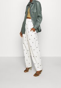 Zelená umělá kožená bunda s límcem, béžový zkrácený top a bílé široké kalhoty s tmavými tečkami, doplněné o sandály na vysokém podpatku.