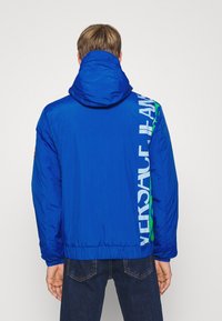 Blå lättviktsjacka med huva, med en stor vit logotyp och gröna accenter längs ena sidan. Slät, glänsande textur med elastisk nederkant.