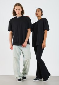 Två modeller bär stora svarta t-shirts med rund halsringning. Den ena bär ljusblå jeans; den andra bär svarta ribbade utsvängda byxor och sneakers.